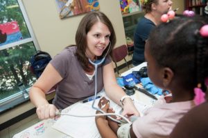 A white woman smiles as she uses a stethoscope & blood pressure cuff to measure a young Black student's blood pressure