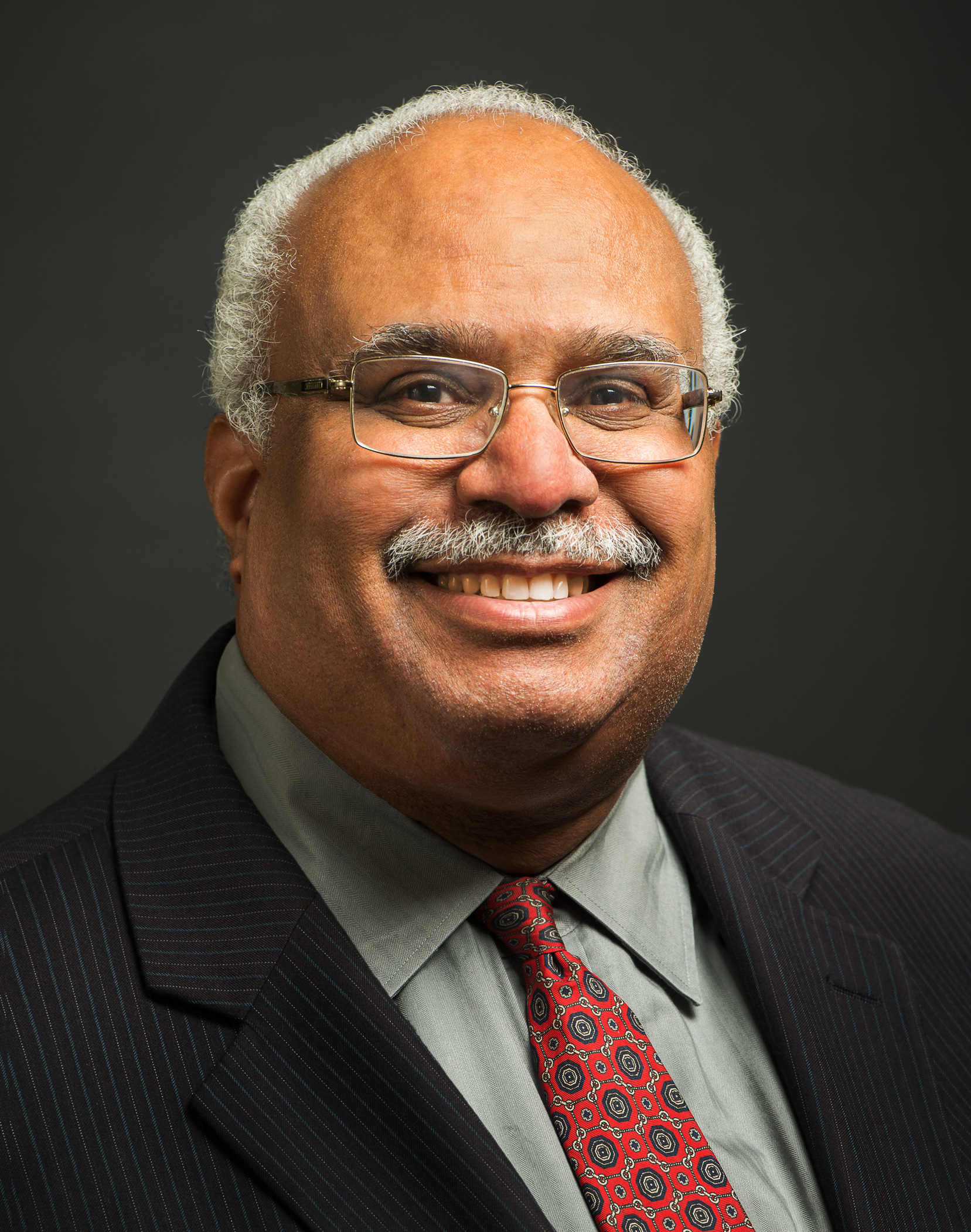 Dr. Georges Benjamin, a Black man with white hair& a white mustache, and glasses smiles. He's wearing a black suit, light green shirt, & red tie.