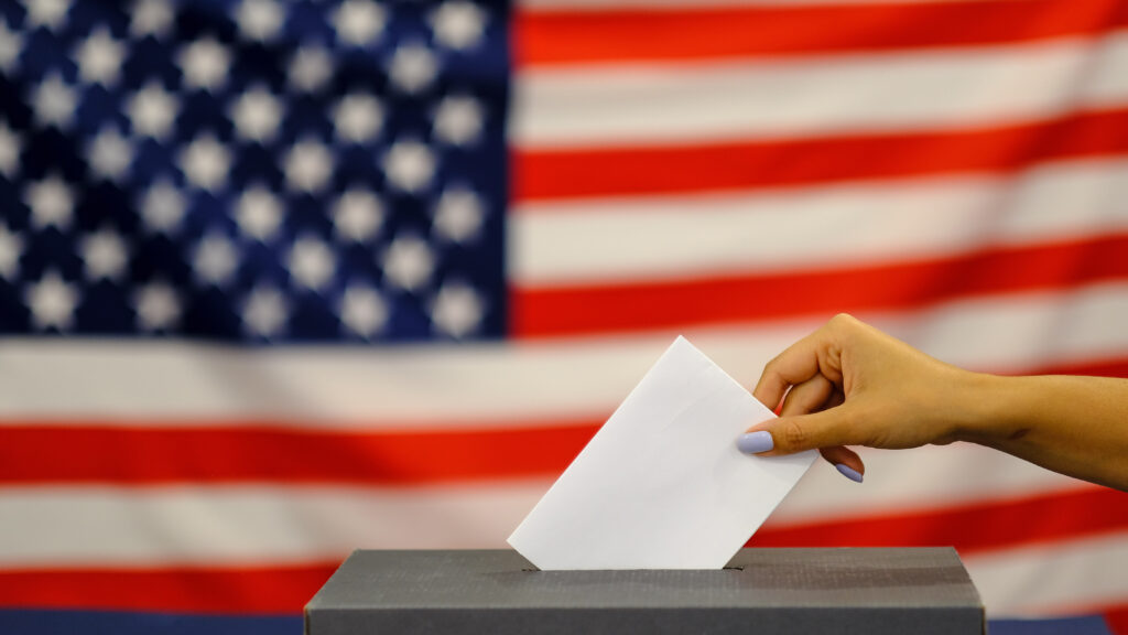 woman putting a ballot in a ballot box on election day. Close up of hand with white votes paper on usa flag background