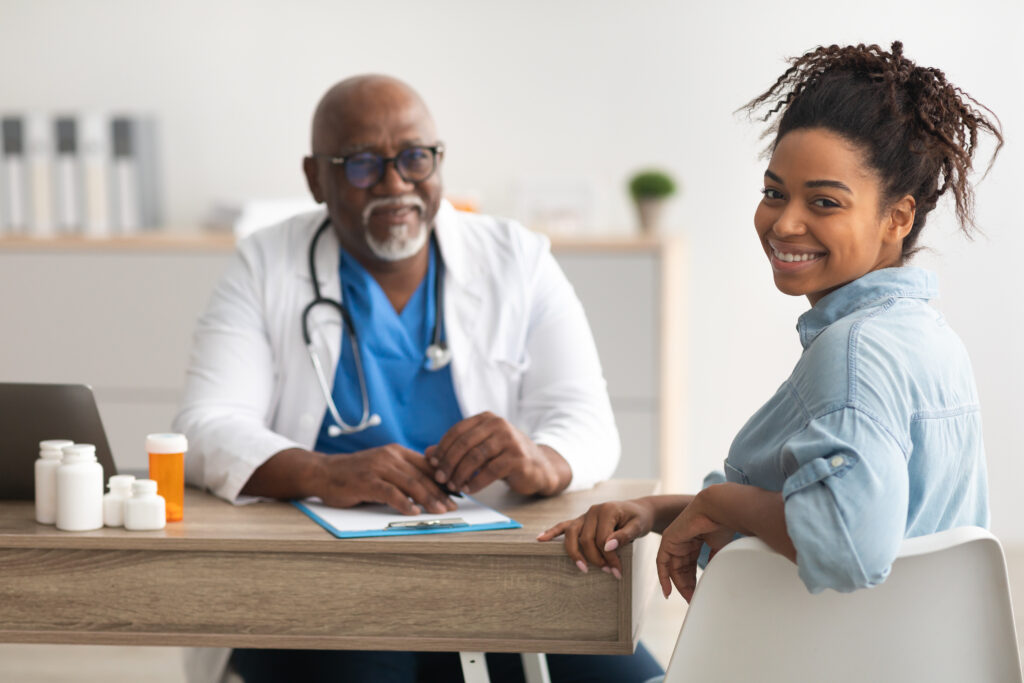 Portrait of smiling African American female patient receiving medical consultation and looking back at camera on foreground, mature male doctor sitting at desk in the blurred background